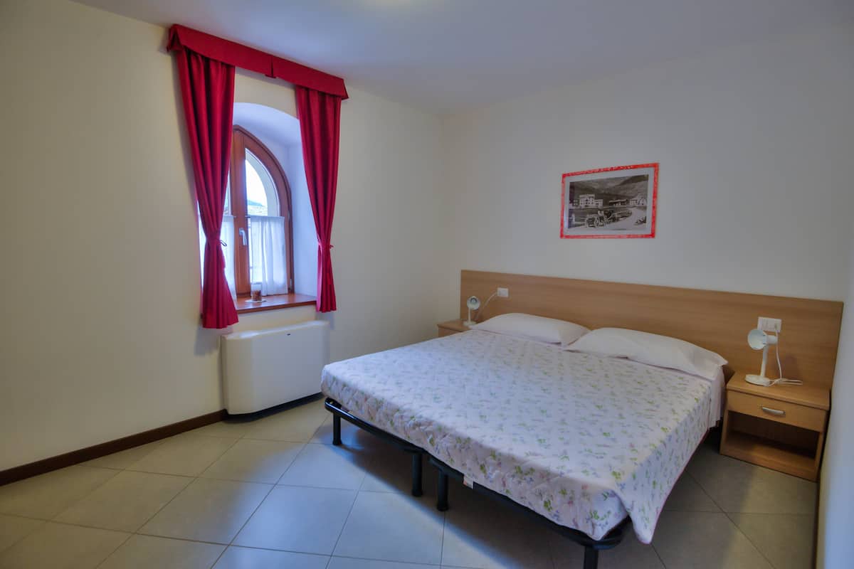Een slaapkamer met een bed, nachtkastje, bedlampje, raam met rode gordijnen, radiator en een ingelijst beeld aan de muur.