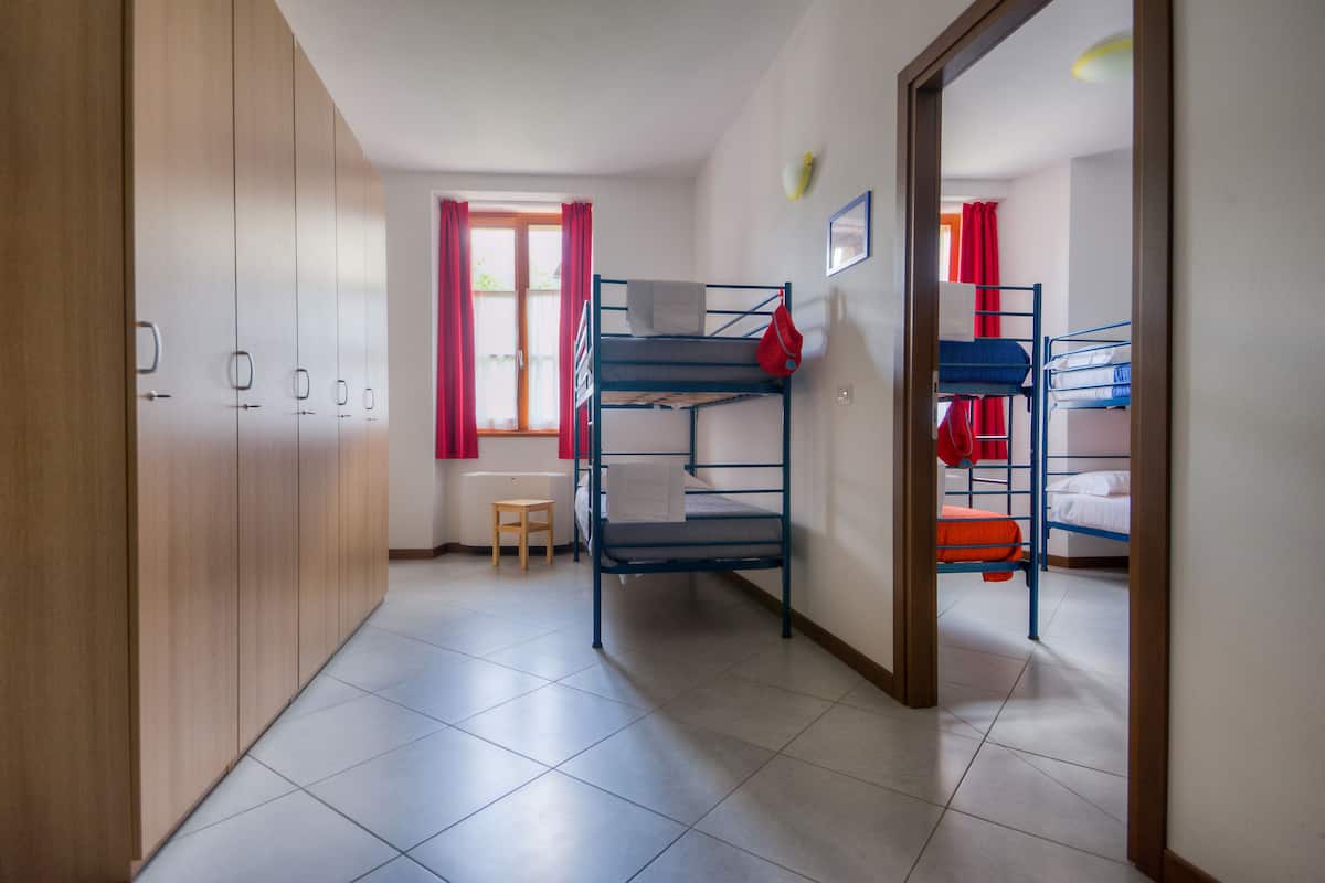 Een slaapzaal met stapelbedden, een houten garderobe, een tafeltje en een raam met rode gordijnen.