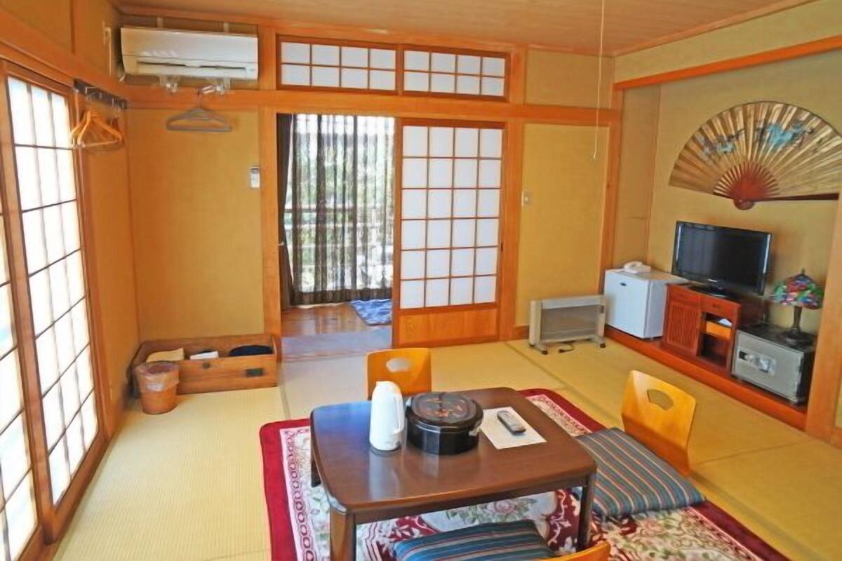 Een traditionele Japanse kamer met tatami vloeren, een lage tafel met een waterkoker, een televisie en een ventilator aan de muur.