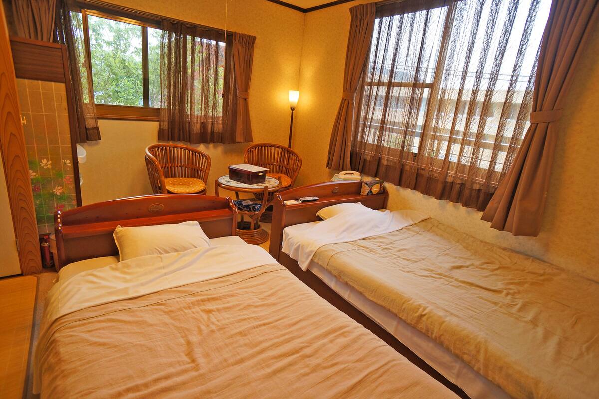 Een hotelkamer met twee eenpersoonsbedden, een houten stoel, een klein tafeltje met een lamp en een raam met gordijnen.