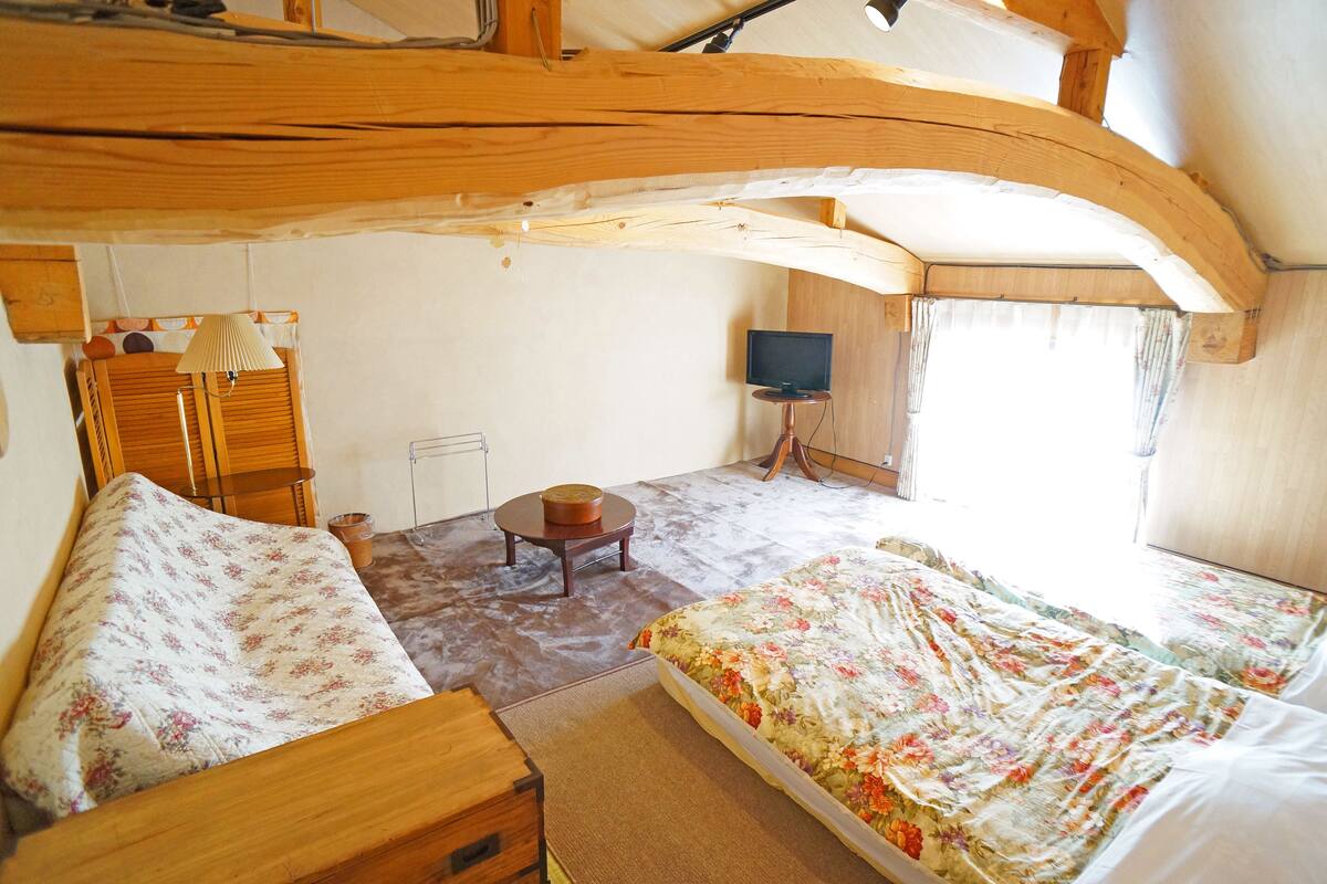 Een kamer met een bed, een houten bankje, een tafeltje, een televisie en een raam met gordijnen.