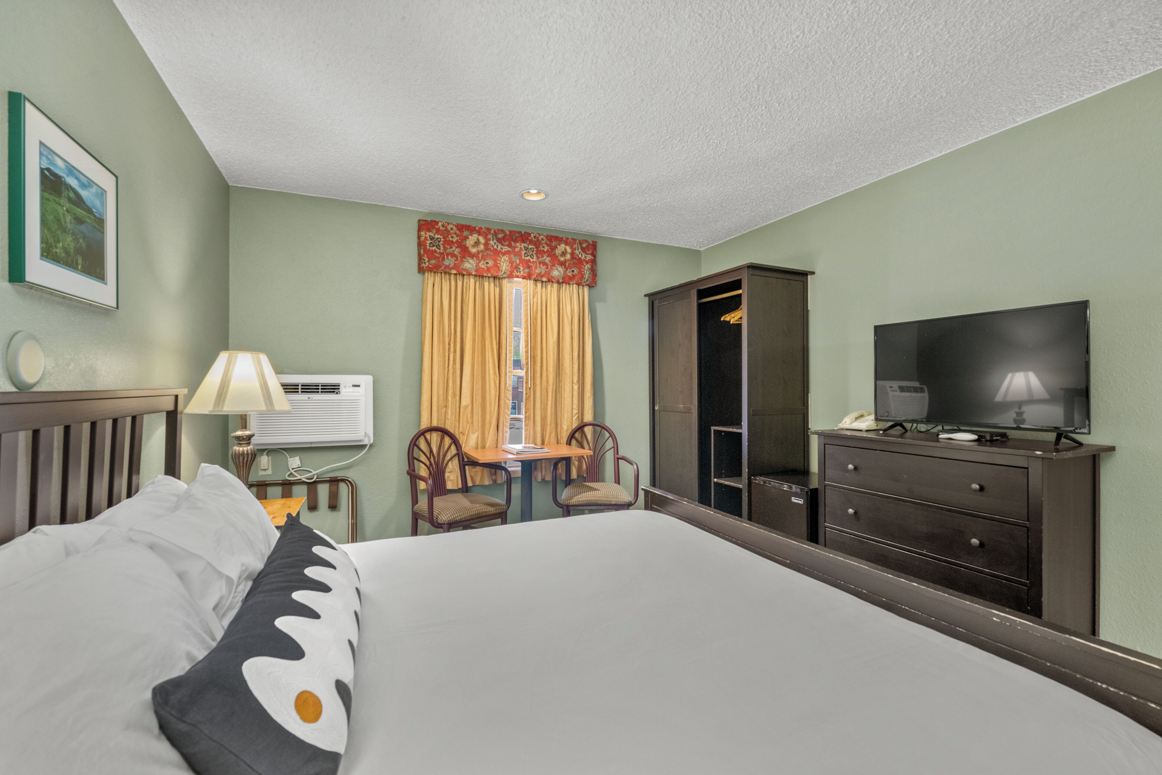 A hotel room with a bed, a desk, two chairs, a television, a wardrobe, and a window with curtains.