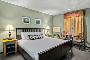 A hotel room with a bed, two bedside lamps, a nightstand, a table with chairs, and a window with curtains.