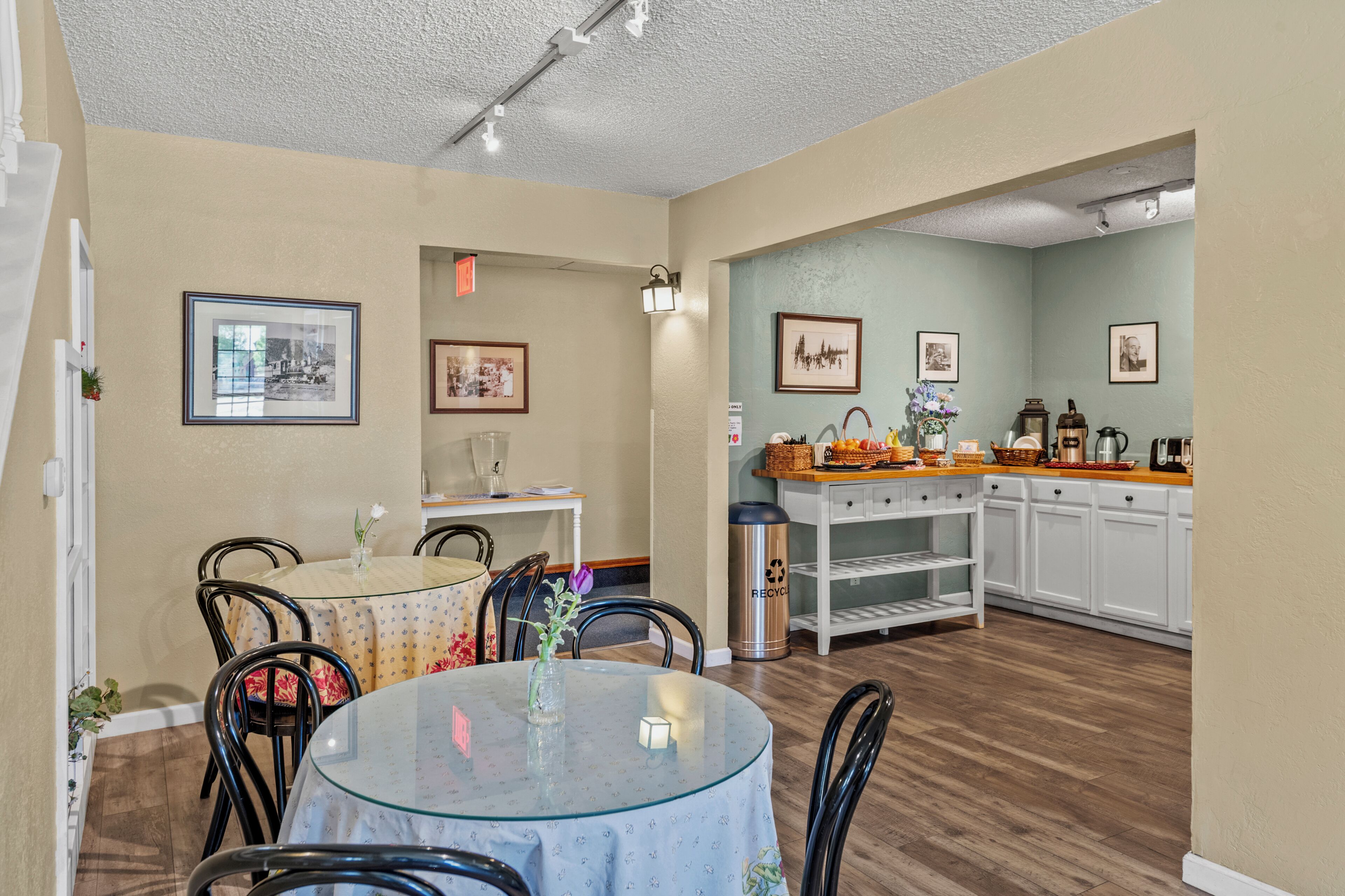 A dining area with a round glass table, chairs, and a vase with flowers. There are framed pictures on the walls and a kitchen area with cabinets and a countertop.
