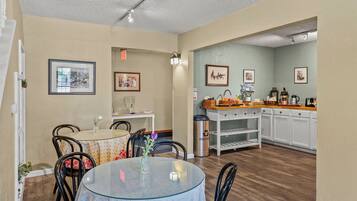 A dining area with a round glass table, chairs, and a vase with flowers. There are framed pictures on the walls and a kitchen area with cabinets and a countertop.