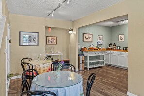 A dining area with a round glass table, chairs, and a vase with flowers. There are framed pictures on the walls and a kitchen area with cabinets and a countertop.