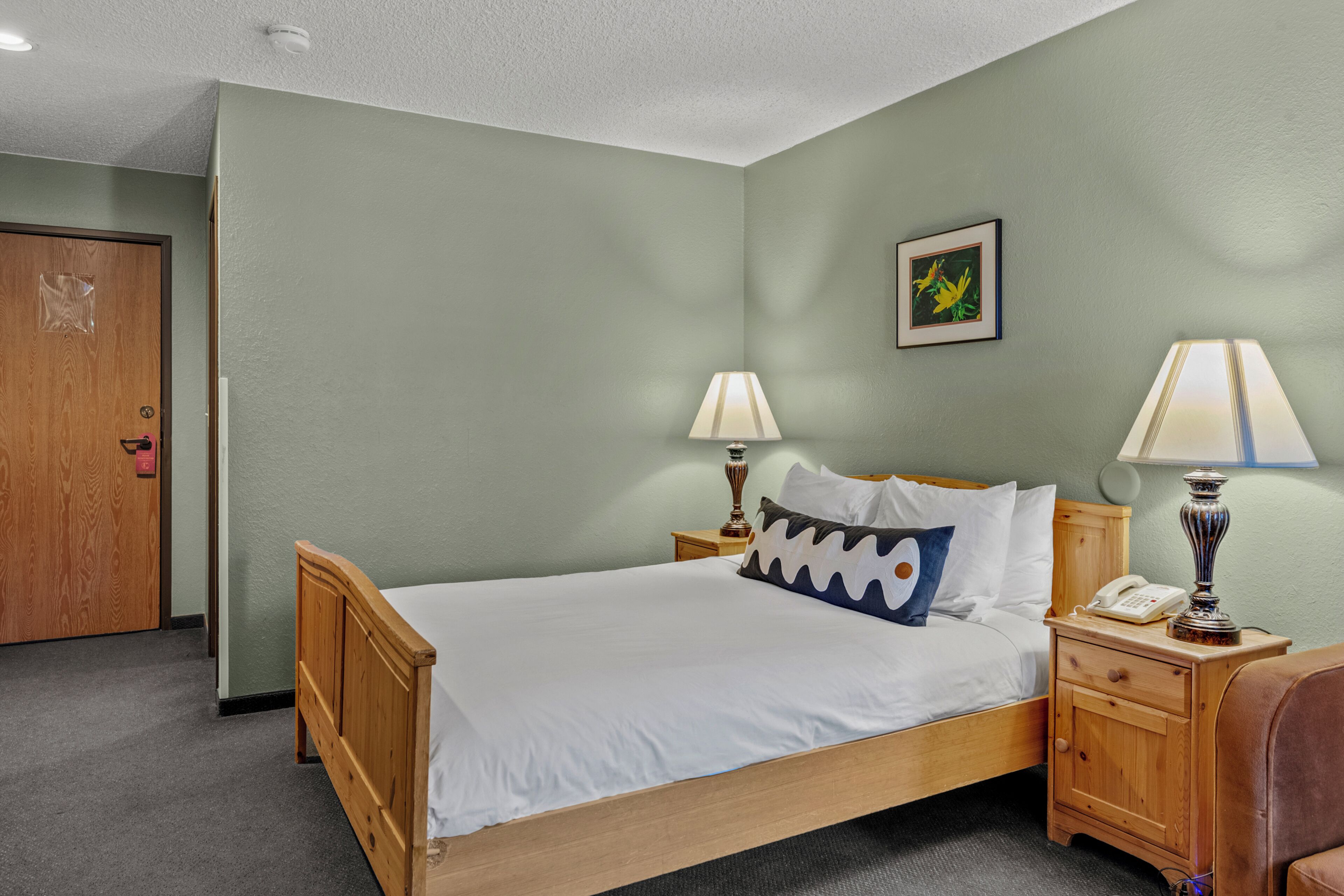 A single-bed room with a wooden headboard, two bedside lamps, a nightstand with a phone, and a door with a red handle.