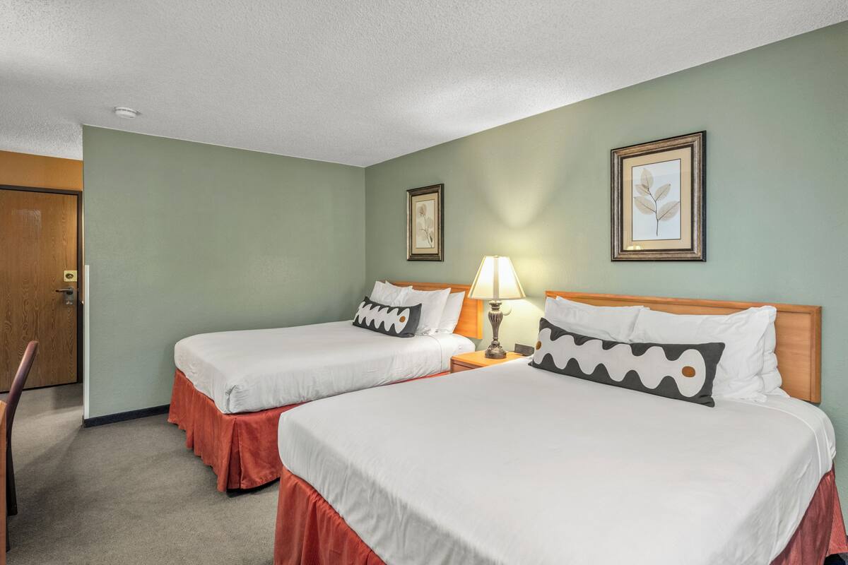 A hotel room with two beds, a wooden door, a framed picture on the wall, and a lamp on the nightstand.