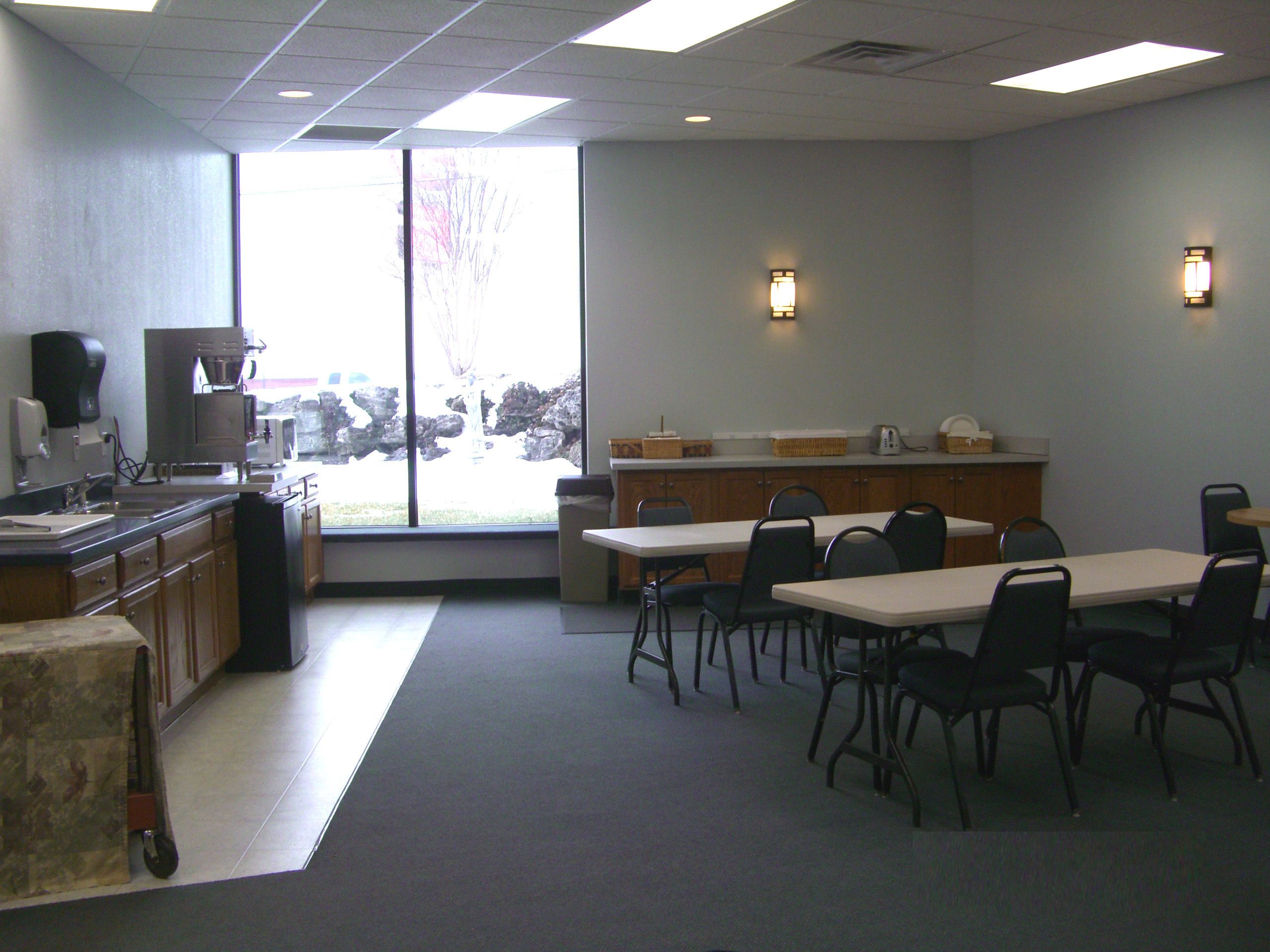 A conference room with a large table and chairs, a kitchenette, and a view through a large window.