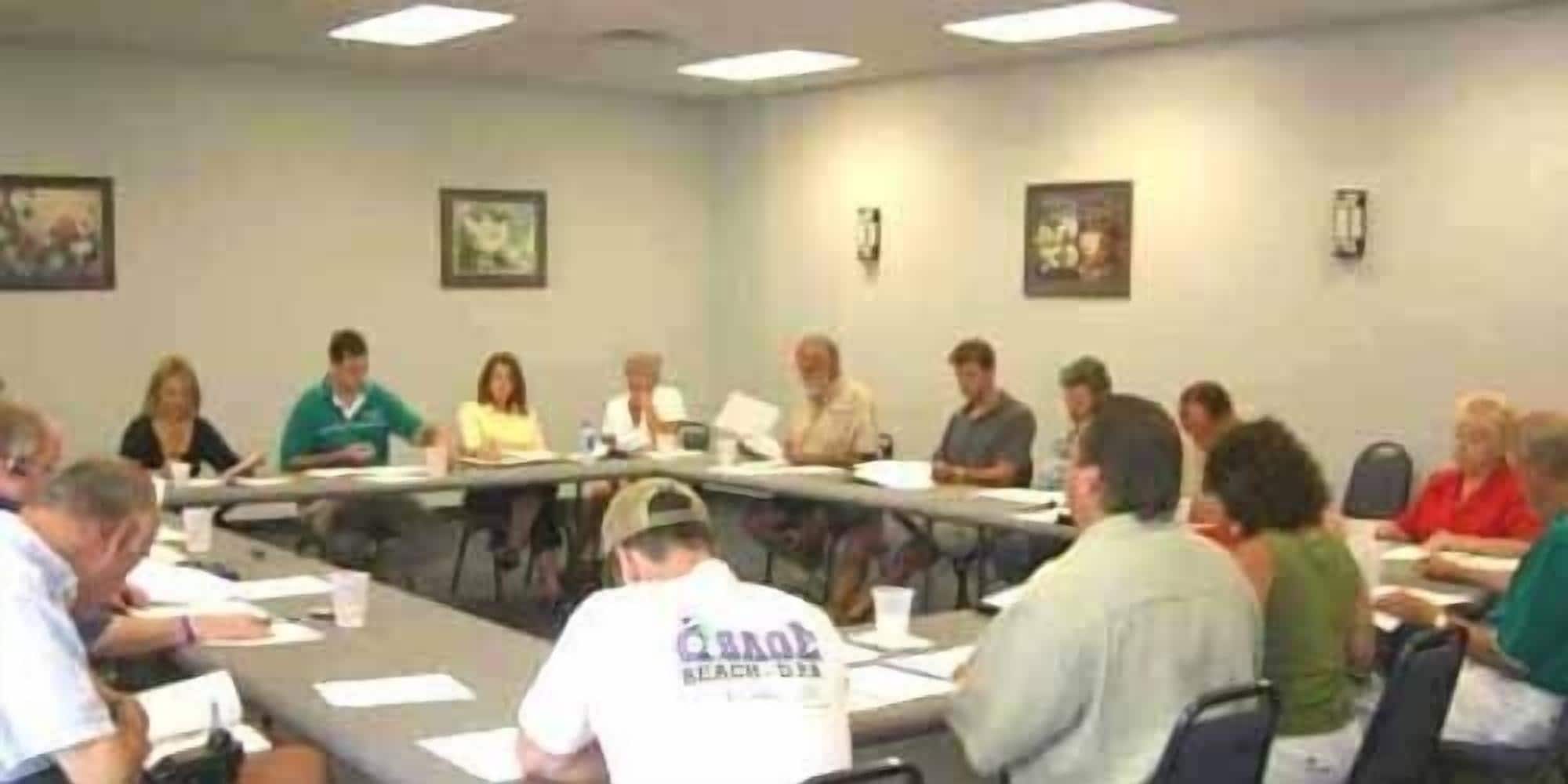 A conference room with a long table surrounded by chairs, papers, and a few individuals seated.
