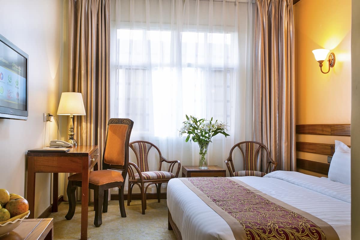 A hotel room with a bed, two chairs, a desk, a lamp, a vase with flowers, and a television.
