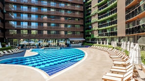 Un espace piscine aménagé avec des chaises longues et des parasols, entouré d’un immeuble à plusieurs étages doté de balcons.