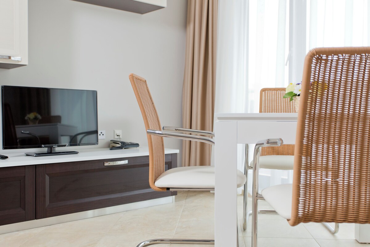 A modern hotel room with a flat-screen TV, a dining area with wicker chairs, and a white table.