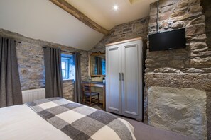 Une chambre avec un mur en pierre, un lit recouvert d’une couverture à carreaux, une armoire, un bureau avec une chaise et un téléviseur fixé au mur.