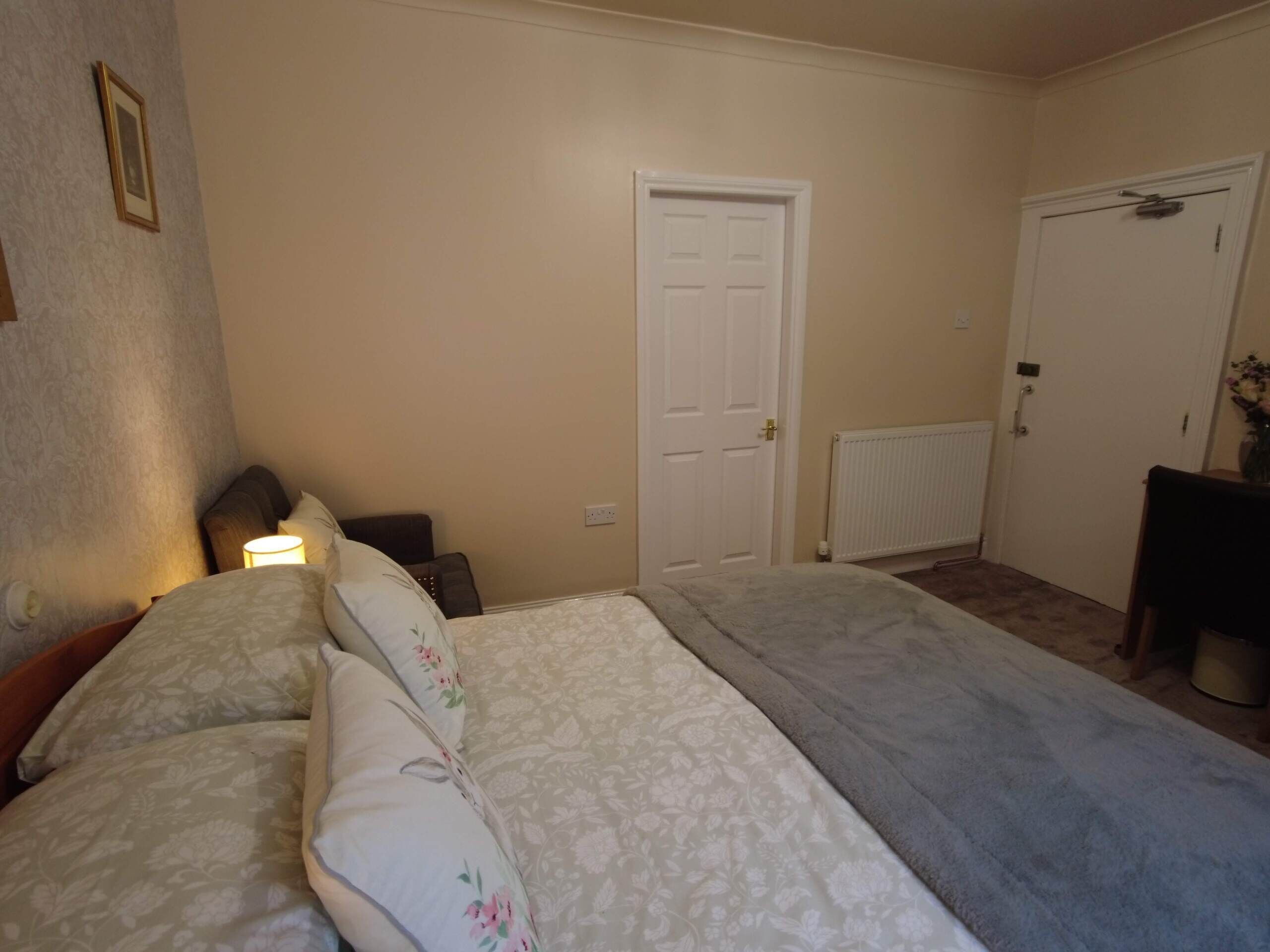A bedroom with two beds, a white door, a radiator, and a small table with a vase.