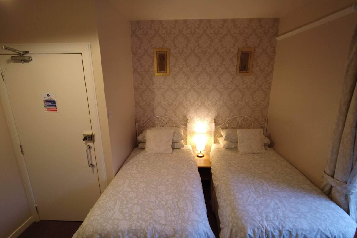 A small, single-room hotel room with two single beds, a door with a fire action sign, and a patterned wallpaper.