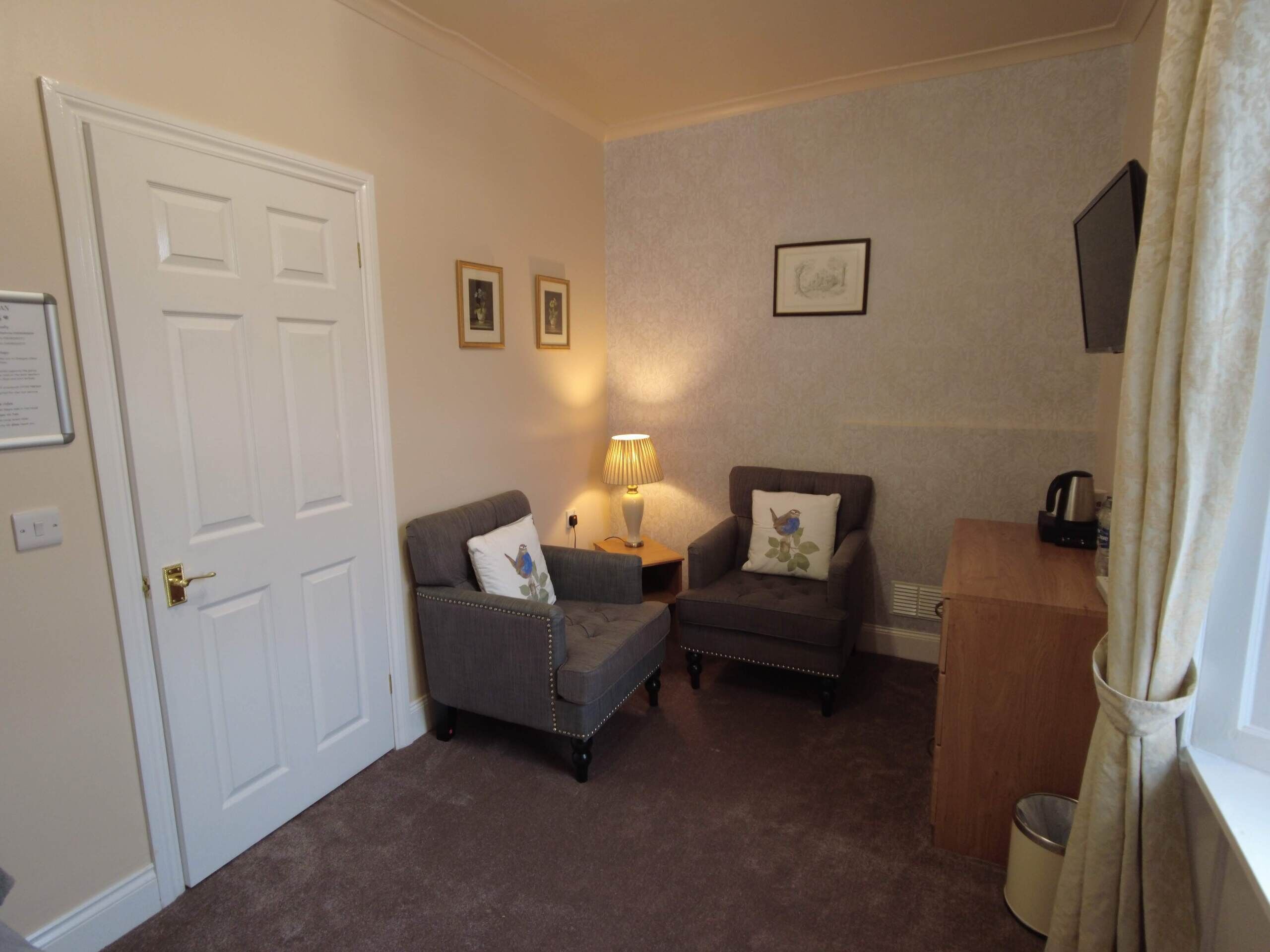 A small, cozy room with two armchairs, a lamp, a wooden side table, and a door.
