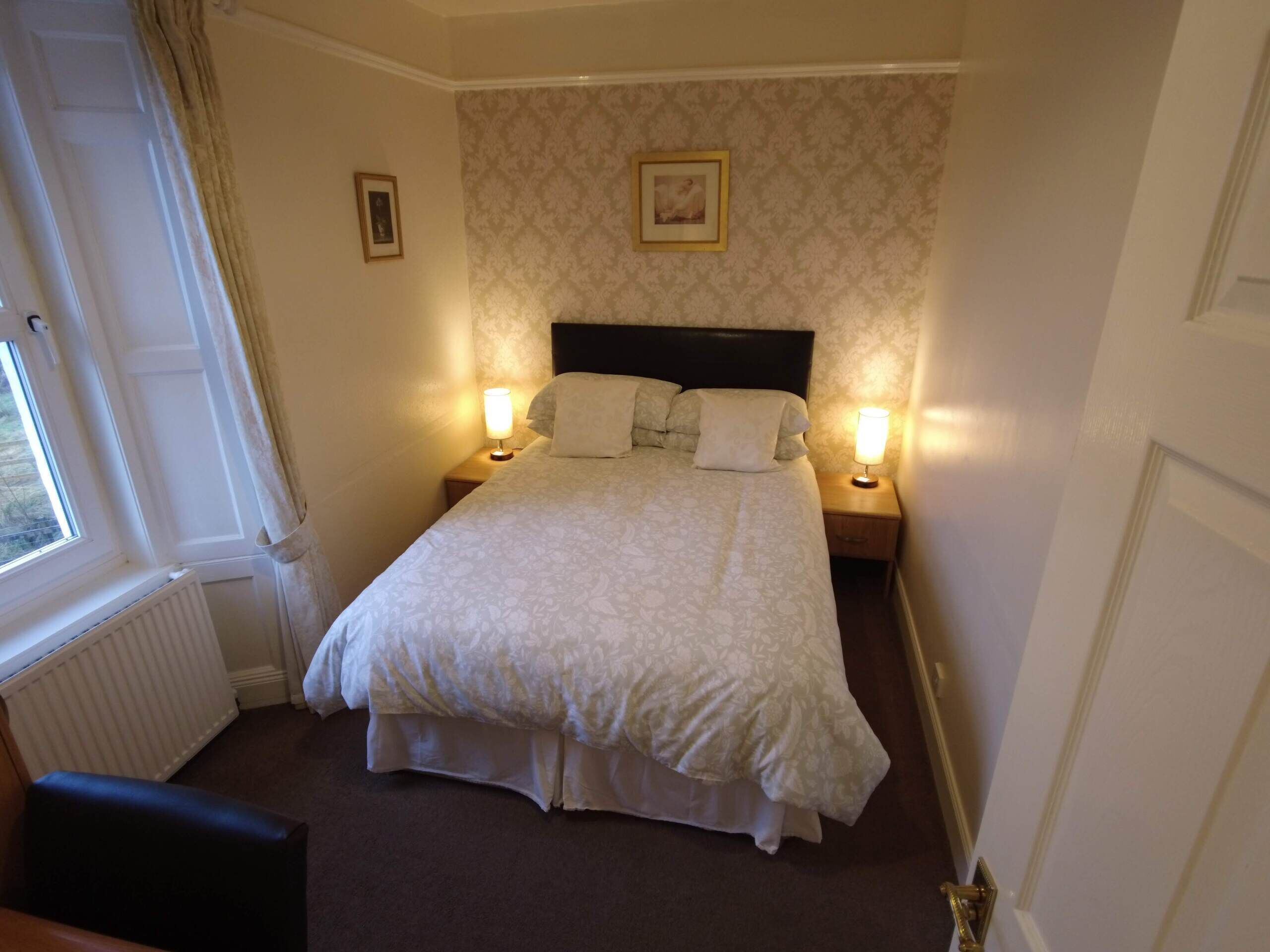 Un lit bien fait, avec une tête de lit, deux tables de chevet avec des lampes, une fenêtre avec des rideaux et un radiateur.