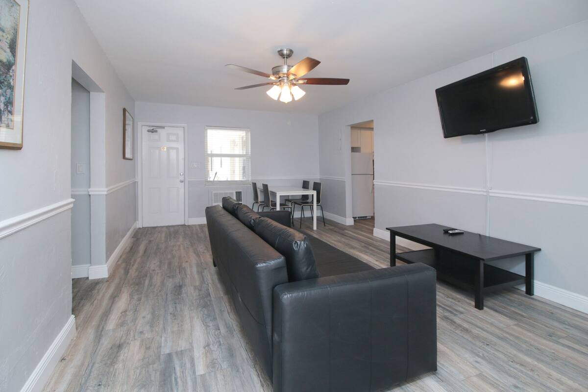 A modern living room with a black leather sofa, a dining table, and a flat-screen TV mounted on the wall.