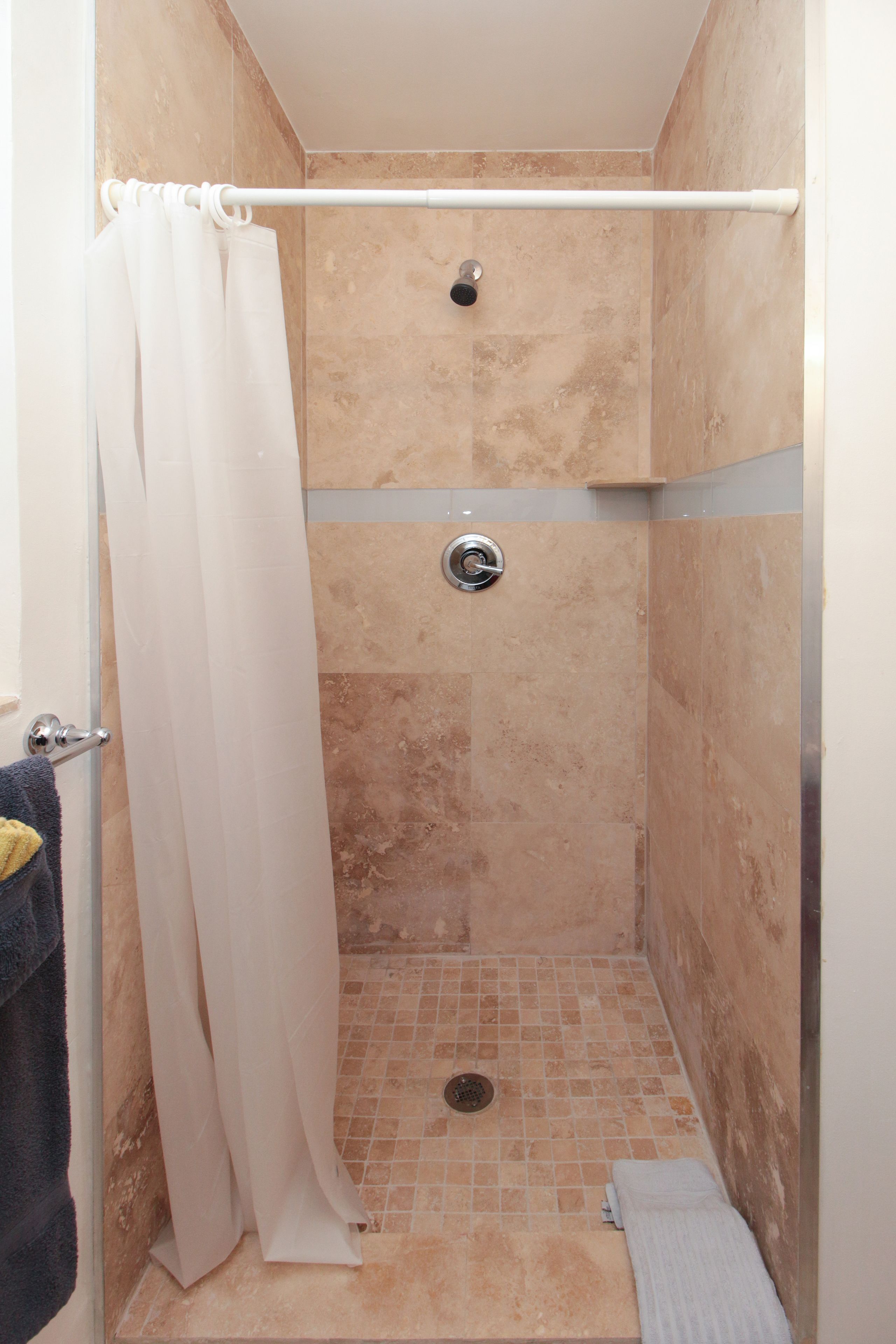 Un pequeño box de ducha con azulejos, una cortina blanca, un perchero y un cabezal de ducha.