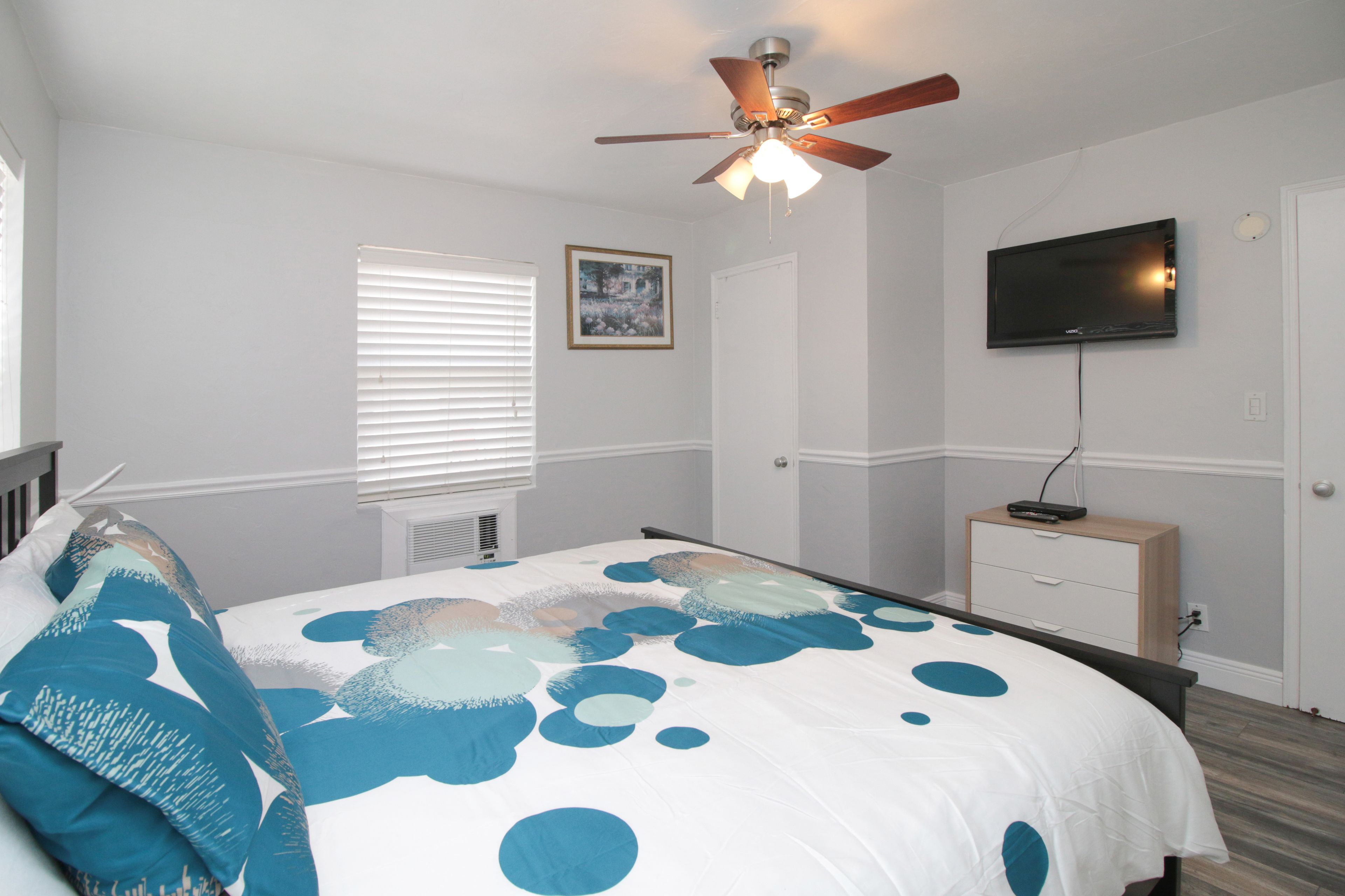 A bedroom with a bed, a ceiling fan, a television, a window with blinds, and a bedside table.