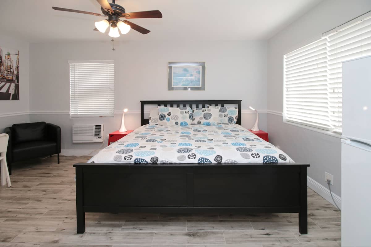 A bedroom with a bed, a ceiling fan, a window with blinds, a bedside table with a lamp, and a wall-mounted air conditioner.