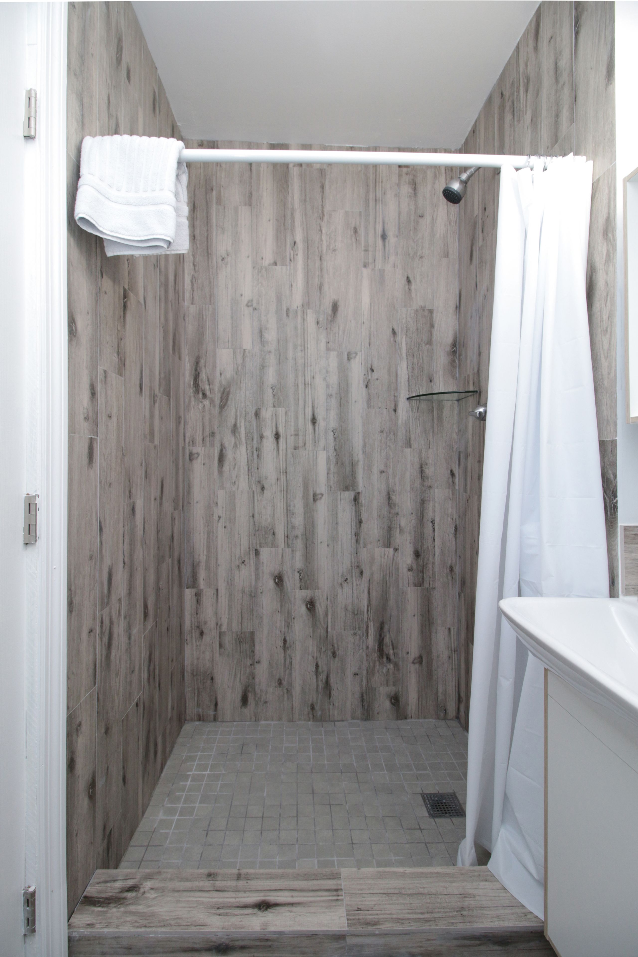 A shower with a wooden wall, a white curtain, and a tiled floor.