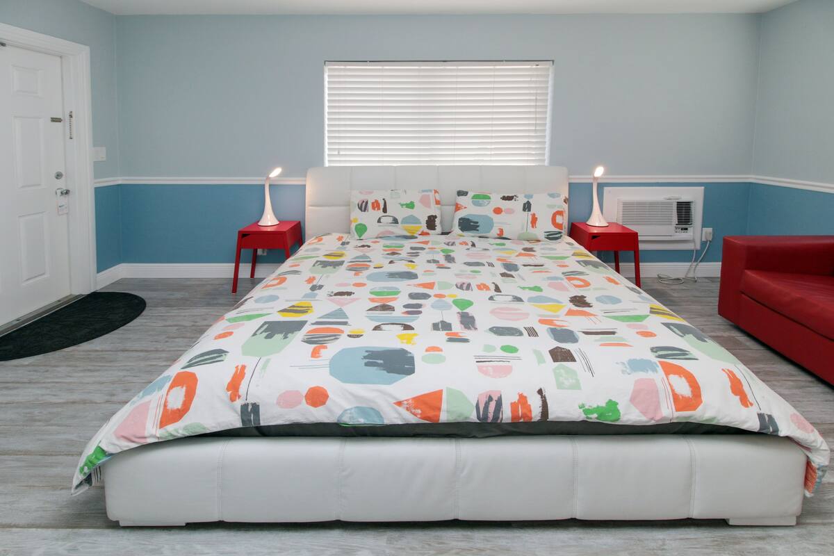 A bedroom with a bed, two nightstands, a red chair, a window with blinds, and an air conditioning unit.