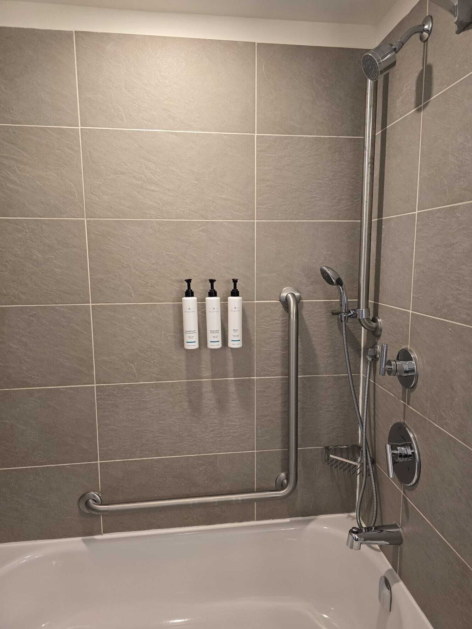 A bathroom with a bathtub, a shower with a handheld showerhead, and three bottles of toiletries on the wall.