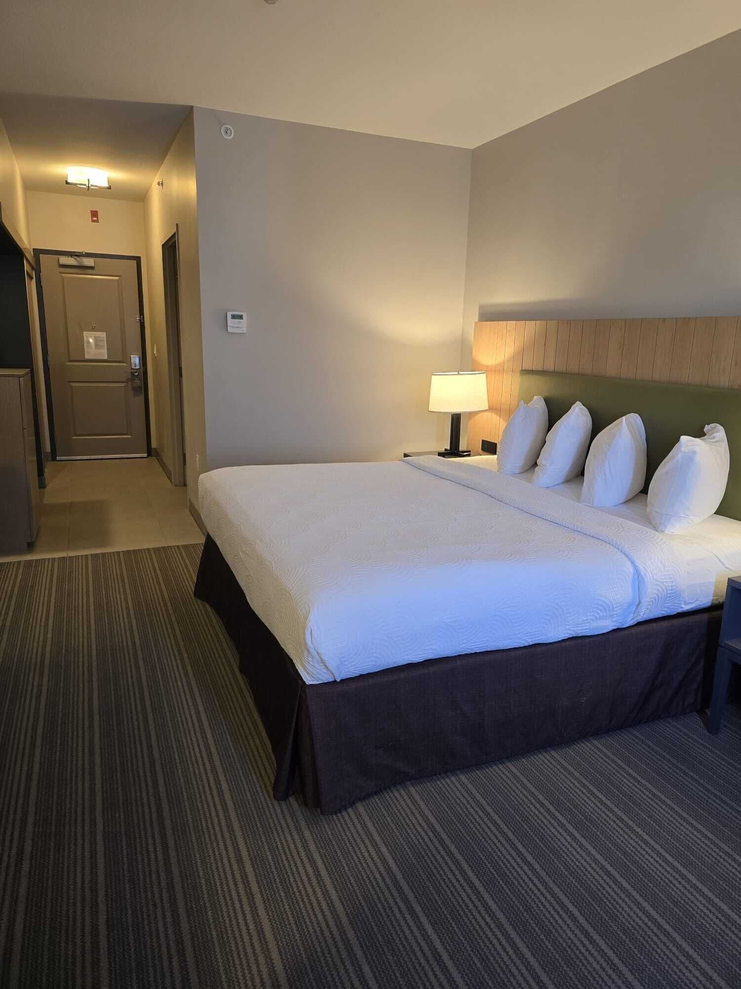 Een hotelkamer met een groot bed, een nachtkastje met lamp, een houten hoofdeinde, een vloerbedekking en een deur naar een gang.