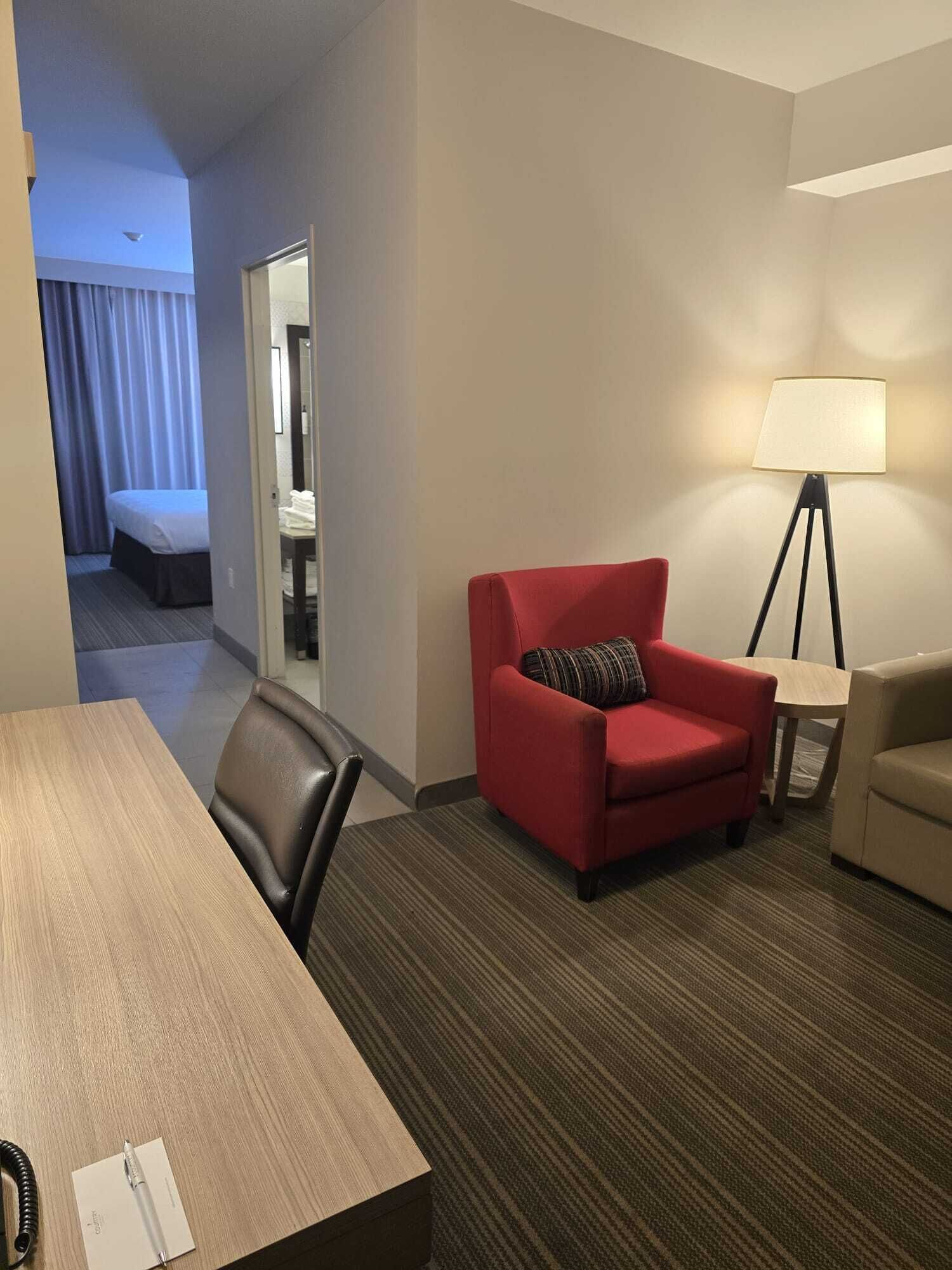Una habitación de hotel con una cama, un sillón rojo, un sillón color beige, una mesita y una lámpara de pie.