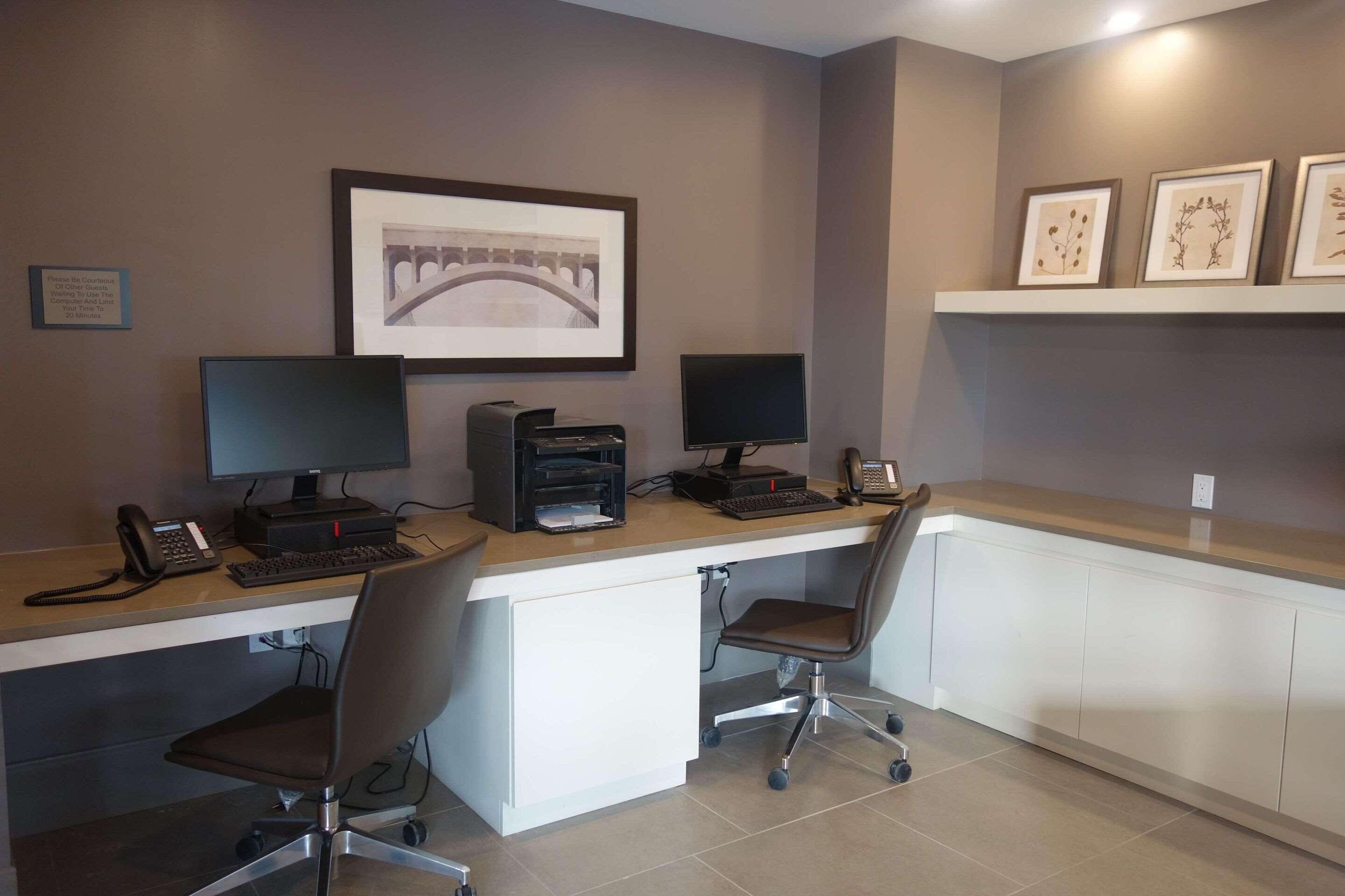 Un espace de bureau moderne comprenant deux bureaux, chacun équipé d’un écran d’ordinateur, d’un clavier et d’un téléphone. Des tableaux encadrés ornent le mur, et une étagère présente d’autres œuvres d’art encadrées.