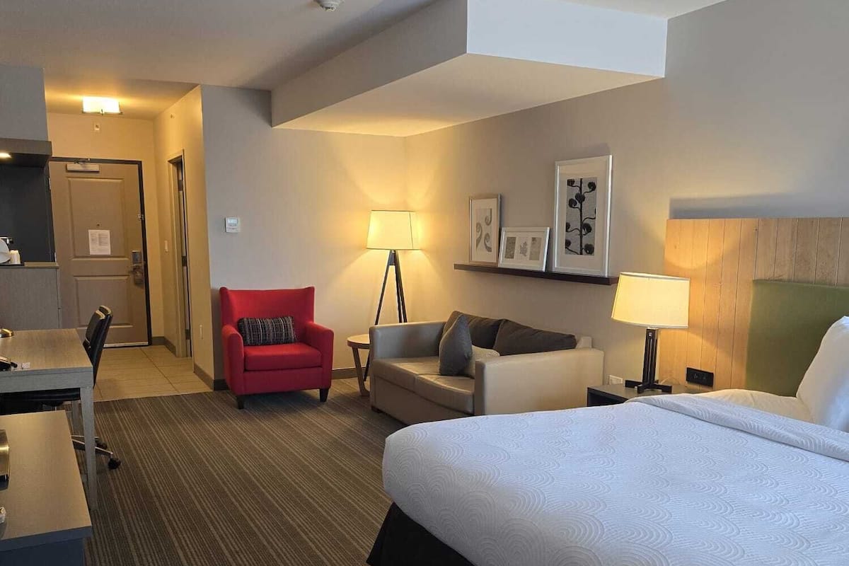 A hotel room with a bed, a desk, a red chair, a sofa, and a lamp.