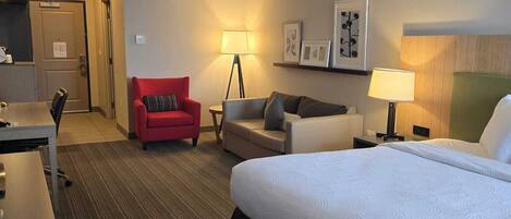 A hotel room with a bed, a desk, a red chair, a sofa, and a lamp.