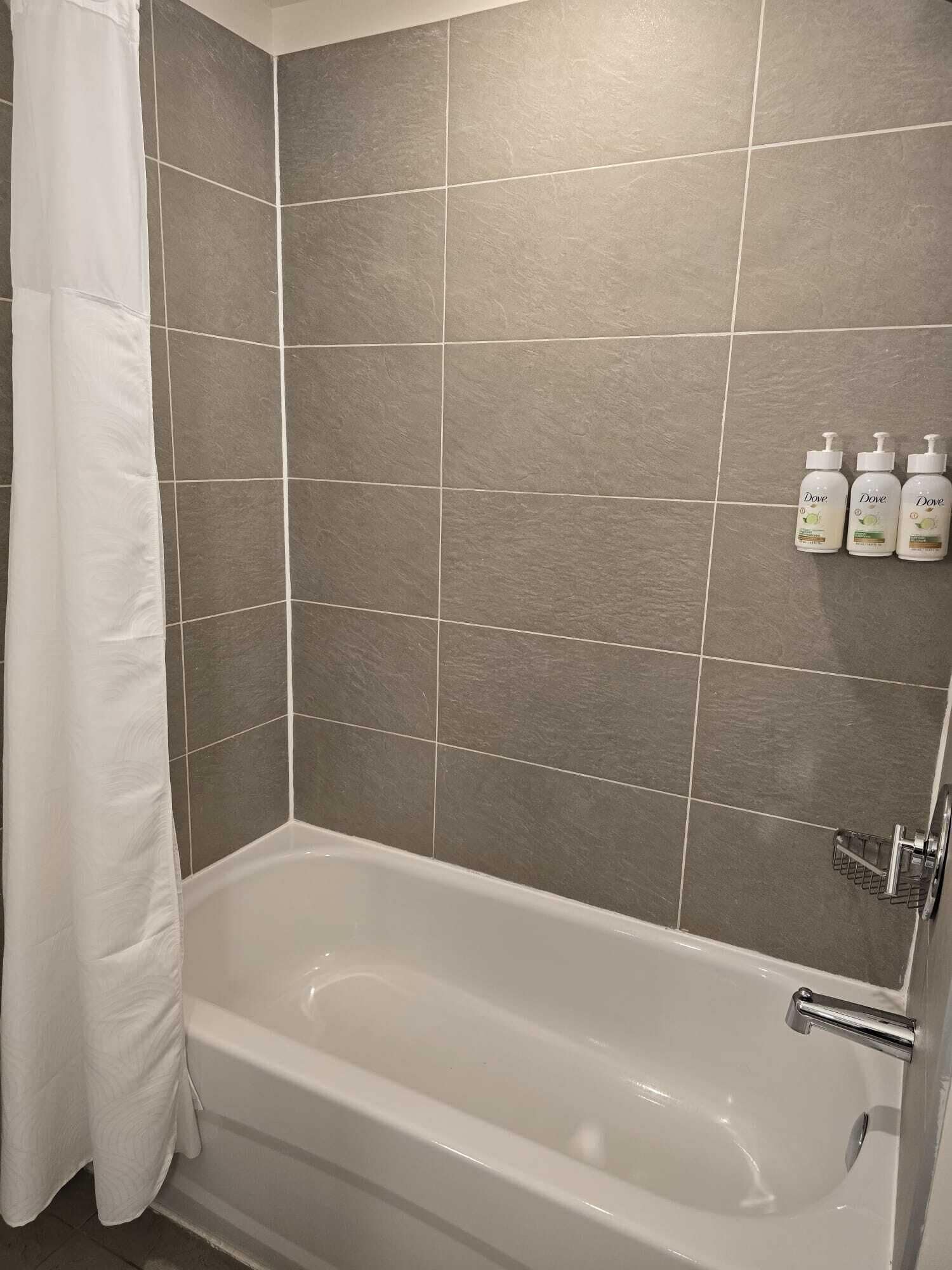 Un baño con bañera, baldosas grises y tres botellas de jabón Dove en la pared.