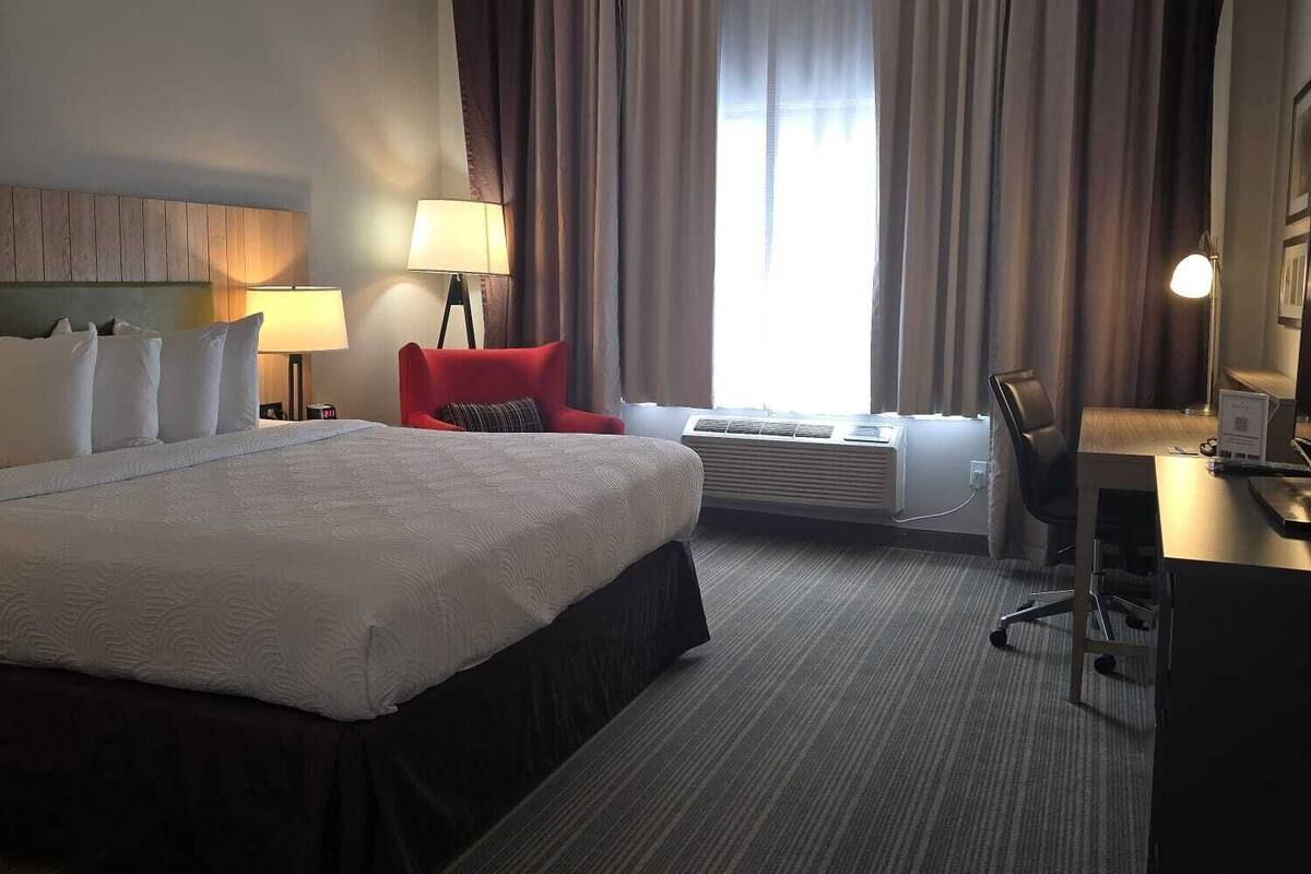 A hotel room with a large bed, a desk, a chair, a lamp, and a window with curtains.