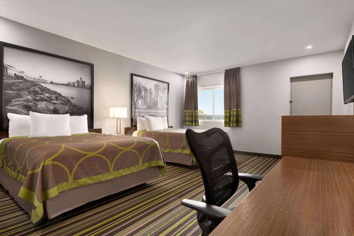 Habitación de hotel con dos camas, un escritorio, una silla, un ventanal con cortinas y obras de arte en las paredes.