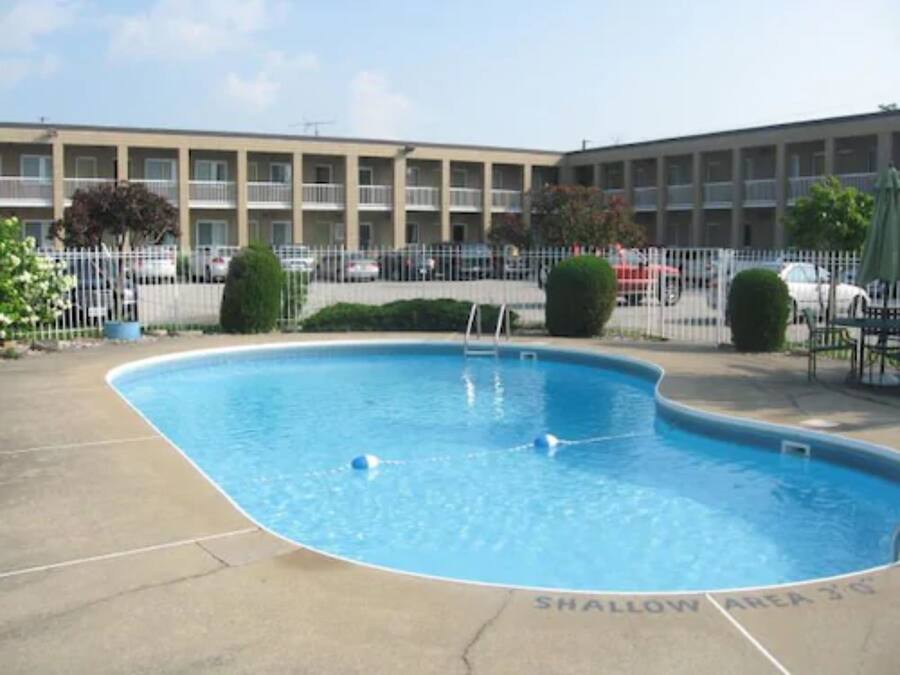 Una piscina con zona poco profunda señalizada, rodeada por una valla, y un edificio al fondo.