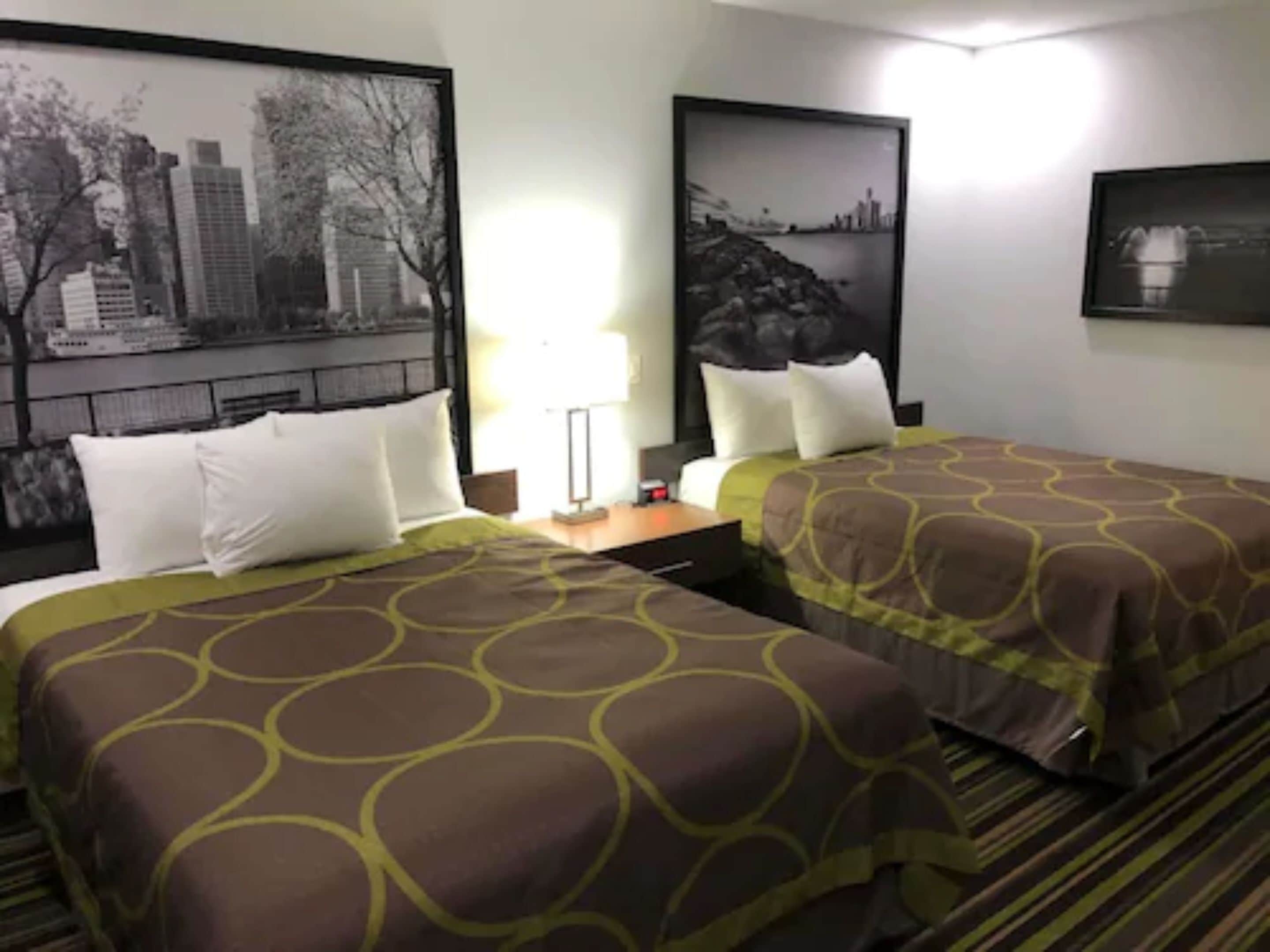 Habitación de hotel con dos camas, una mesita de noche, una lámpara y fotografías enmarcadas de la ciudad en las paredes.