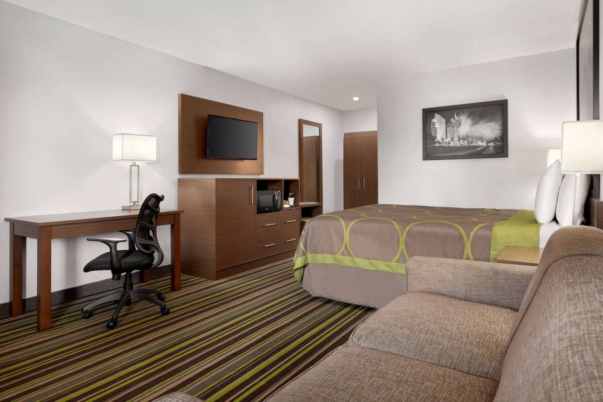 Una habitación de hotel con cama, escritorio, silla, televisión, espejo y un cuadro en la pared.
