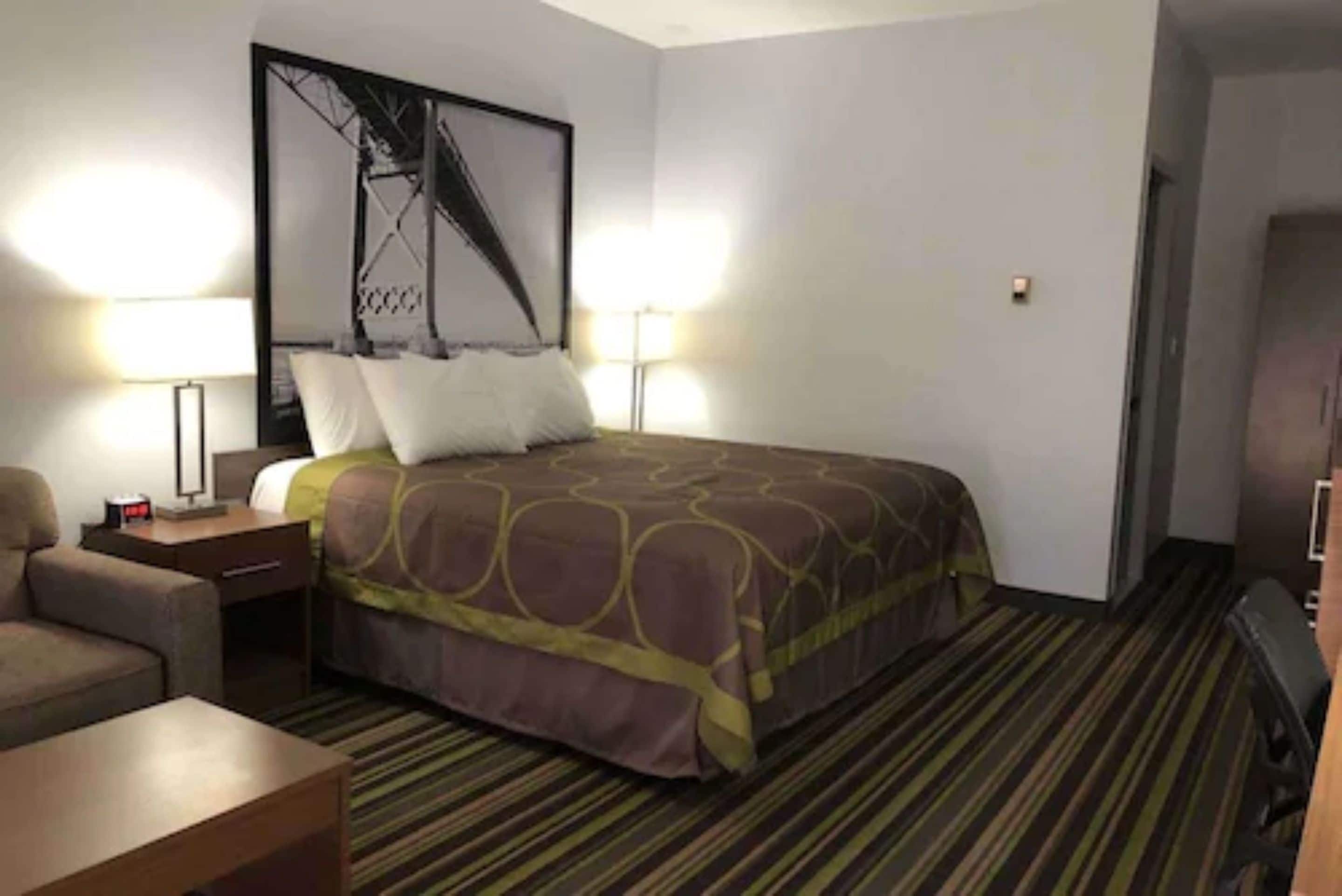 Habitació d'hotel amb un llit, dues làmpades de nit, una cadira, un escriptori i un quadre gran amb marc a la paret.