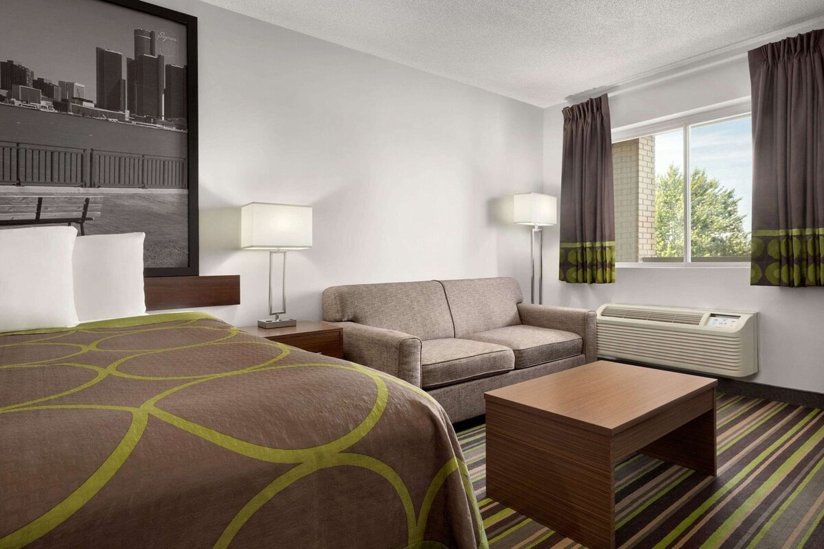 A hotel room with a bed, a sofa, a table, a lamp, a window with curtains, and a wall-mounted air conditioner.