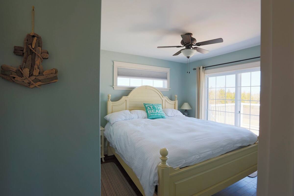 A bedroom with a bed, a ceiling fan, a window with blinds, and a wall-mounted light fixture.