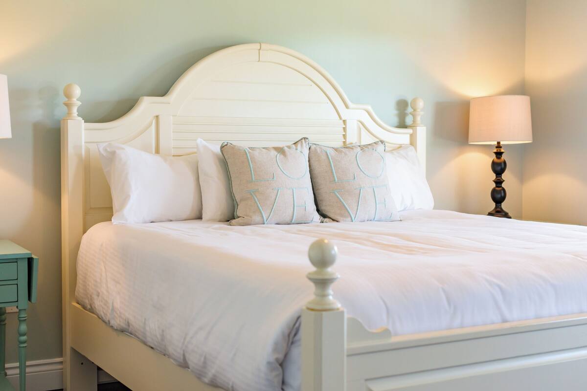 A neatly made bed with white bedding and decorative pillows, a bedside table, and a lamp.