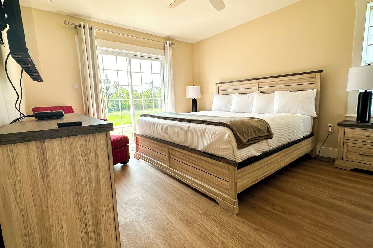 A bedroom with a bed, bedside table, lamp, and a view of the outdoors through a window.