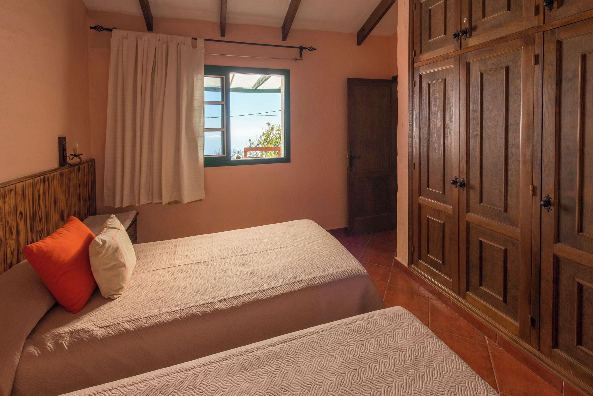 A bedroom with two beds, a window with curtains, a wooden wardrobe, and a wooden headboard.