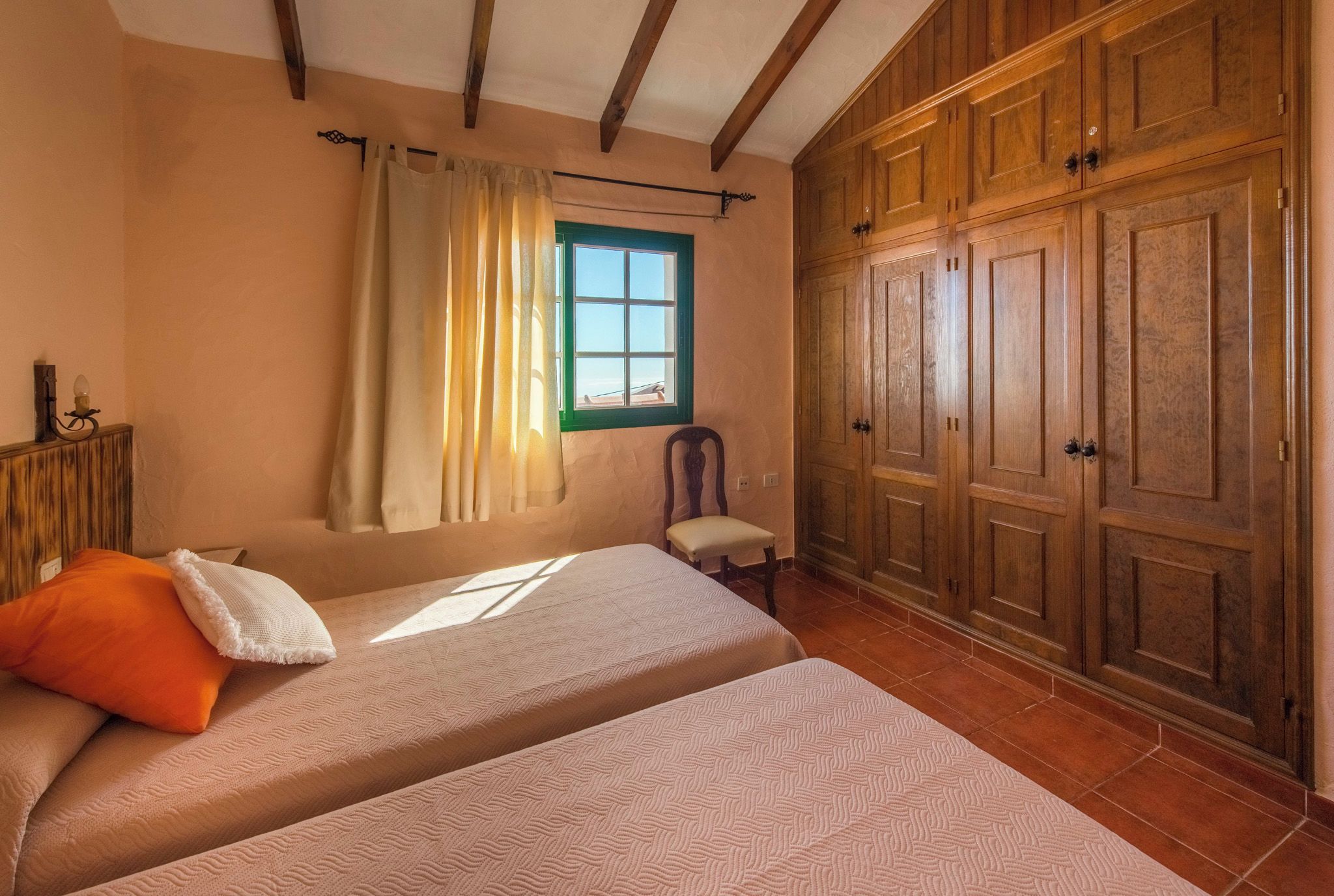 Une chambre avec deux lits, une fenêtre avec des rideaux, une armoire en bois et un petit tabouret.