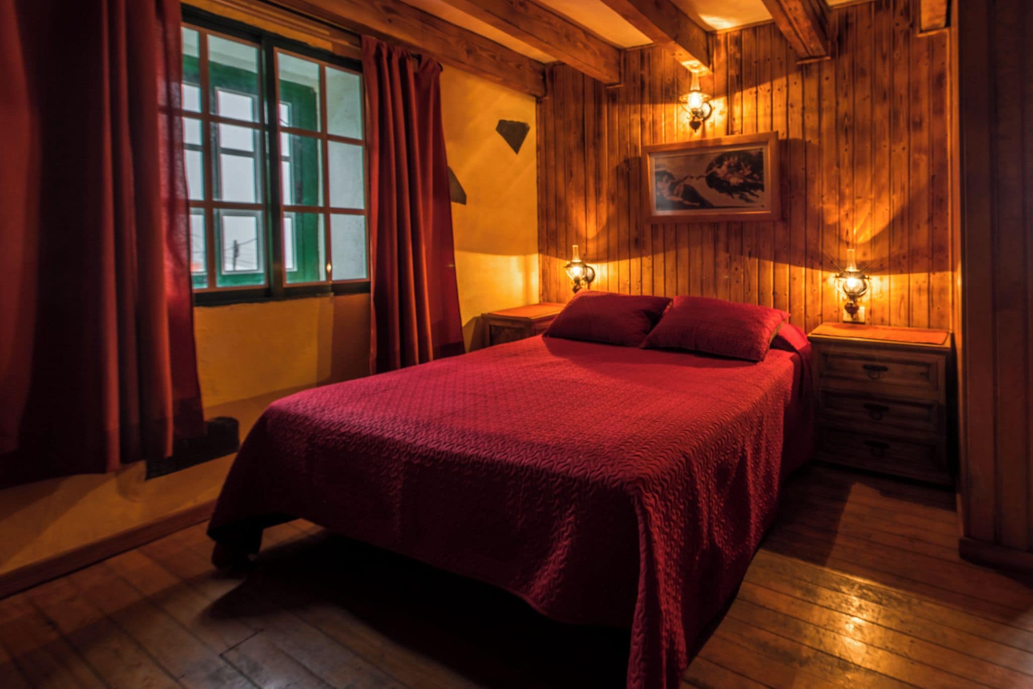 Una habitación de madera con una cama, dos mesitas de noche, un ventanal con cortinas y una lámpara fijada a la pared.