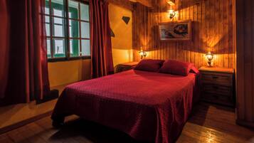 Una habitación de madera con una cama, dos mesitas de noche, un ventanal con cortinas y una lámpara fijada a la pared.