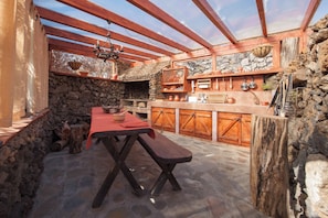 A rustic outdoor kitchen with a stone wall, wooden beams, and a wooden table with a bench.